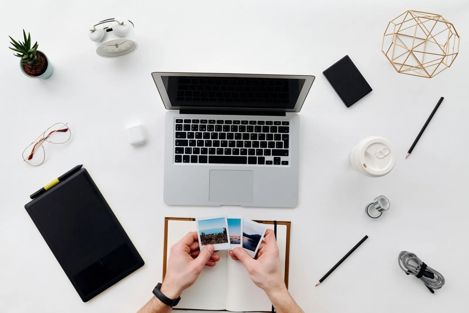 Overhead photo of a white desk with a laptop, a drawing tablet, a notebook, and hands holding three instant photo prints.